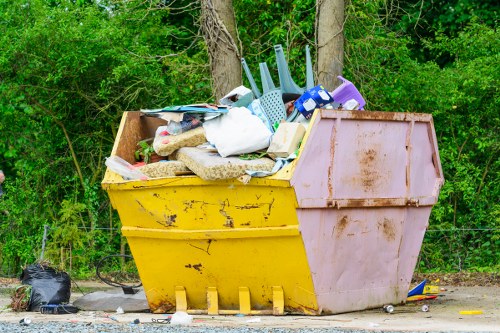 A large, yellow and pink skip filled with a mixture of household rubbish and outdoor waste, including plastic containers, cardboard boxes, fabric items, and a mop, positioned on a gravel surface in front of a lush green backdrop of trees and bushes. The skip appears weathered with visible rust and dirt, and some waste extends slightly over the top edge. Surrounding the skip are small black plastic bags and scattered debris, indicating a recent clearance activity. The scene suggests an outdoor rubbish removal process that might be handled by a service like Office Clearance Camden Town, specialising in comprehensive waste disposal in the Camden Town area of London. The natural surroundings and urban environment denote a typical setting for domestic or small-scale commercial rubbish collection in this part of central London.