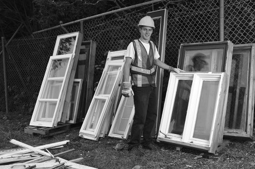 A male worker in a high-visibility safety vest and a white hard hat stands outdoors on a grassy area near a chain-link fence, surrounded by several large, white, double-glazed UPVC window frames leaning against the fence and placed on wooden pallets. The frames vary in size and are made of PVC with metal reinforcements seen along the edges. The worker is smiling and holding one of the window frames, indicating an active rubbish removal or clearance process. In the foreground, additional window components and some loose wooden battens are scattered on the ground. The scene suggests a site for office or residential clearance, with the presence of multiple window frames ready for disposal or recycling, typical of waste management services provided by companies such as Office Clearance Camden Town. The background features a natural environment with trees and a metal fence, implying the clearance is taking place in an outdoor setting, possibly in Camden Town or nearby areas within the postcode region.