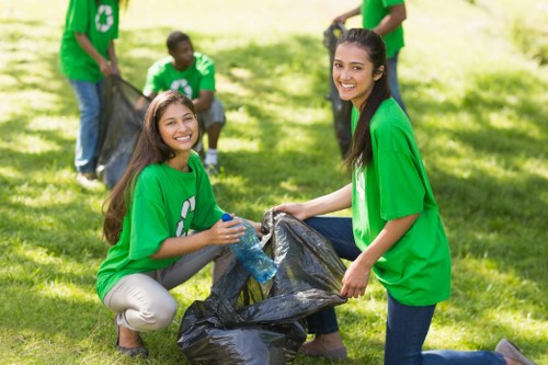 Four young individuals wearing bright green t-shirts with recycling symbols participate in outdoor rubbish collection in a grassy park setting with trees in the background. One woman is kneeling on the grass, smiling, and holding open a large black plastic rubbish bag while a man kneeling beside her also holds the bag open. Behind them, another woman and two men are visible; one man is standing and holding a filled black rubbish bag, while the other appears to be placing waste into the bag. The volunteers' clothing contrasts with the natural environment, emphasizing their involvement in community clean-up activities. The scene features natural sunlight, casting soft shadows on the grass, and the focus is on the collaborative effort of waste collection, aligning with general rubbish removal services and community environmental initiatives in Camden Town.