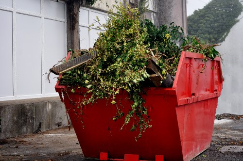 A large, bright red industrial dumpster filled with assorted garden waste and debris is positioned on a paved surface, likely outside a building in Camden Town. The contents include a dense cluster of leafy green branches and stems, along with some small wooden planks and weathered garden tools that are visible among the foliage. The surface of the waste appears rough and natural, with textures of leaves, twigs, and unfinished wood contrasting against the smooth, painted exterior of the dumpster. In the background, there is a white paneled wall or window structure and a concrete base, with some ivy or climbing plants partially visible, adding to the outdoor setting. The lighting suggests a natural daylight environment, illuminating the scene evenly and highlighting the vibrant green of the garden waste against the red container. This image relates to rubbish removal services, such as those offered by Office Clearance Camden Town, which handle garden waste clearance in the local area.