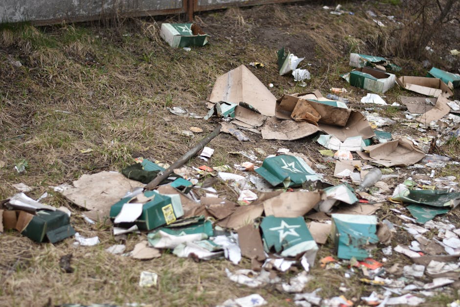 A patch of ground alongside a grassy embankment is littered with discarded cardboard boxes and packaging materials, some flattened and torn, with a mix of brown and green cardboard with white and yell