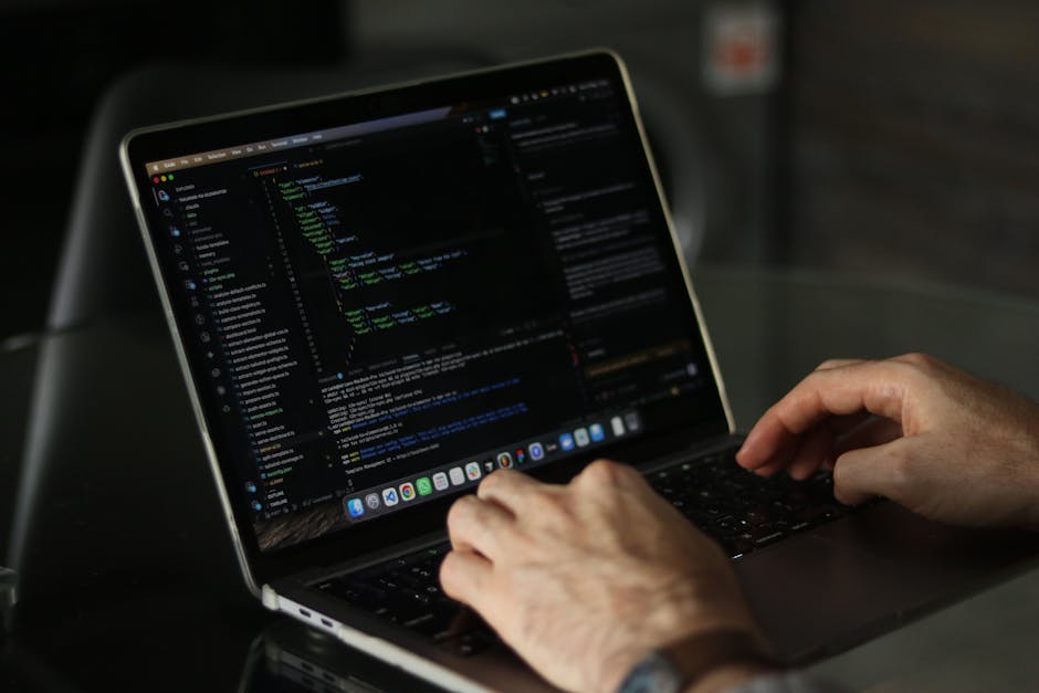 A person using a laptop with a dark-colored exterior and a high-resolution display, showing lines of colorful code on a black background. The individual’s hands are positioned over the keyboard, one
