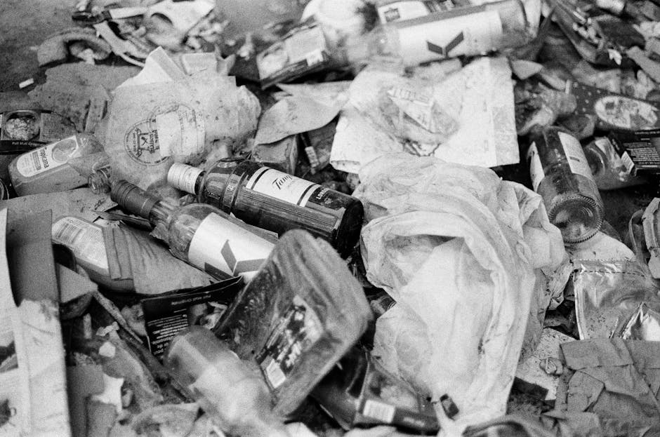 A black wheeled rubbish bin labeled 'St. John's' positioned on a city sidewalk at night, with the lid open and various waste materials and brown paper bags visible inside. The bin is situated close to