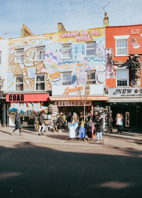 A busy street scene in Camden with a row of multi-story buildings, one featuring a large, colorful mural with graffiti-style artwork and text, including phrases like 'Graffiti for the People' and 'Lon