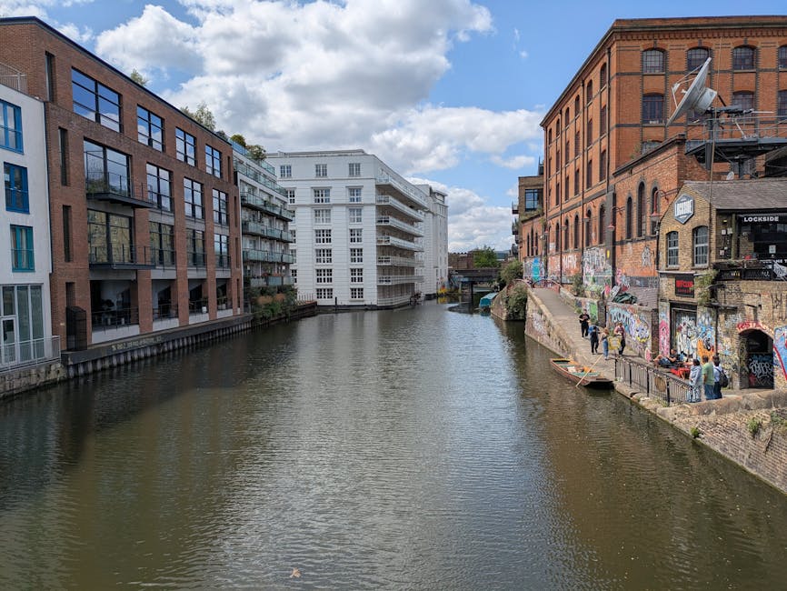 A narrow canal flanked by residential buildings on both sides, with some modern multi-storey apartments featuring balconies and large windows on the left, painted in shades of yellow and white, and ol