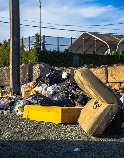 A pile of black rubbish bags and discarded cardboard boxes stacked in front of a red metal door with graffiti and signage, located on an exterior wall of a building with a textured beige surface. The